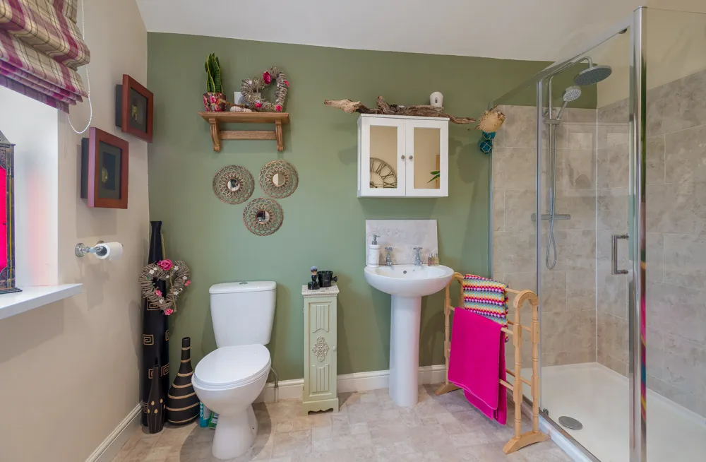 Modern bathroom with white toilet, pedestal sink, glass shower enclosure, and green accent wall with decorative wreaths and a shelf.