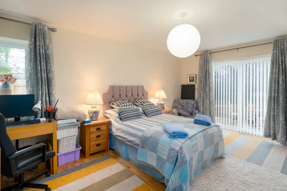 Cozy bedroom with striped bed, patterned chair, wooden desk with computer, floral curtains, and soft lighting.