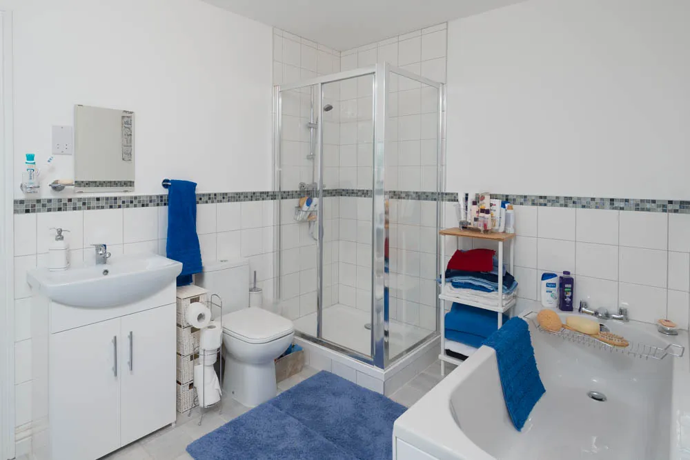 Clean bathroom with white tiles, a shower enclosure, white sink and toilet, blue towels, and a small shelving unit holding toiletries.