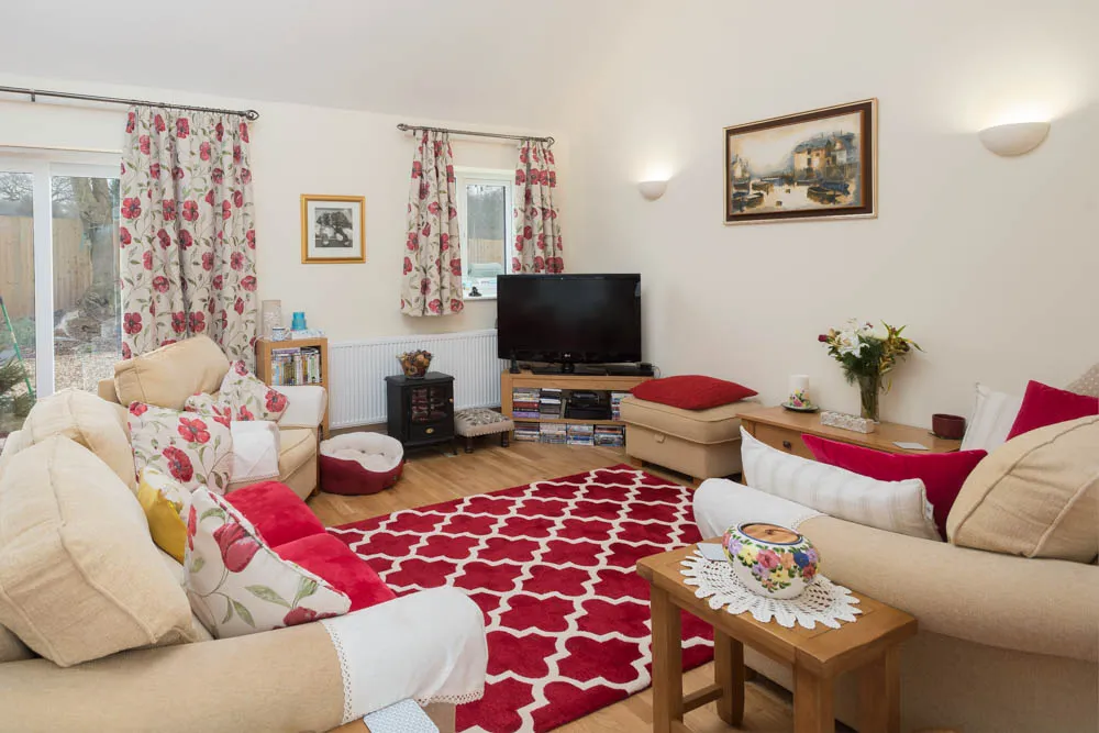 Cozy living room with beige sofas, red and floral cushions, red patterned rug, a TV on a wooden stand, and floral curtains.