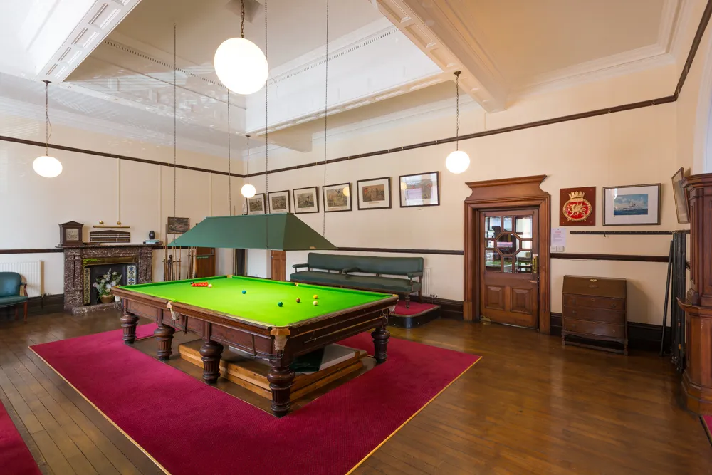 A spacious vintage snooker room with a green baize table, hanging lamps, wooden floor, red carpets, framed pictures, and seating area.