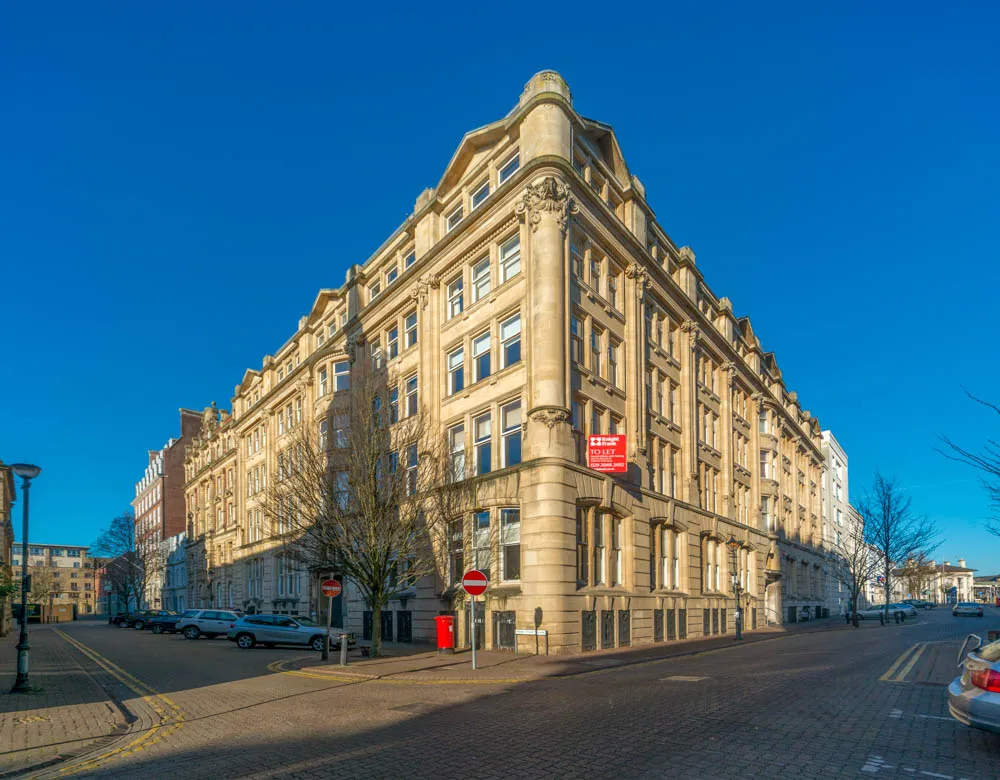 Wide-angle view of a historic beige stone building on a sunny day with clear blue sky and parked cars along the streets.