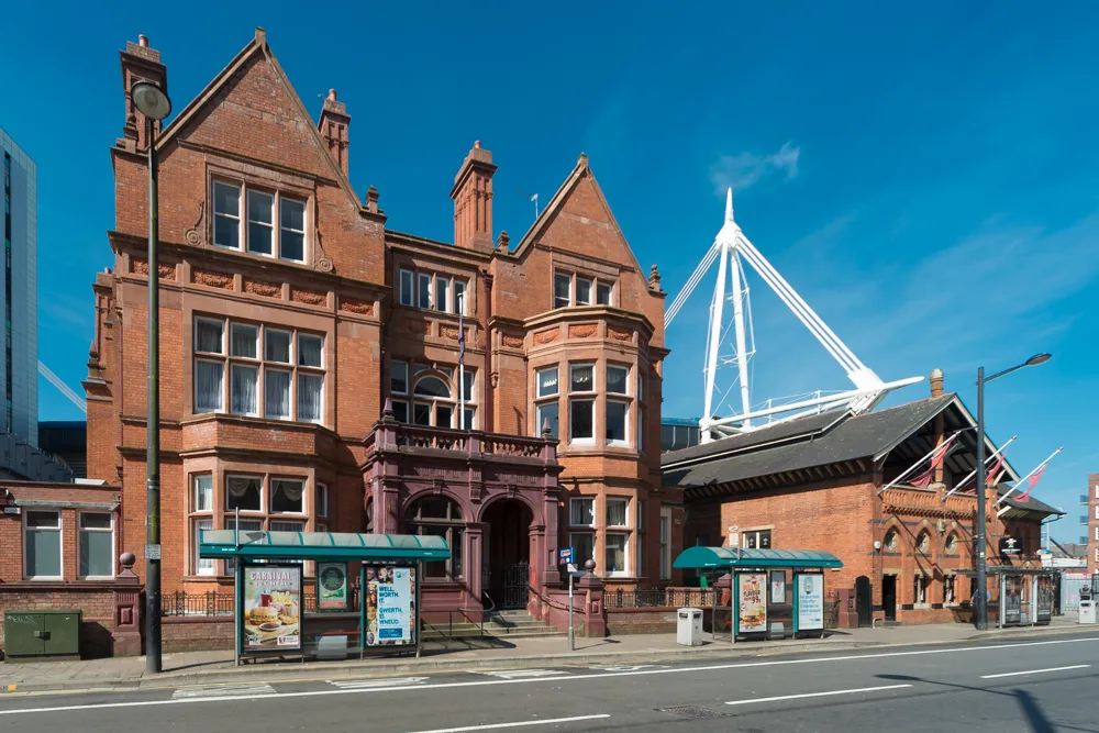 Red brick historic building with bay windows beside a modern white stadium structure under a clear blue sky.