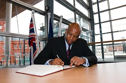 Man in a suit signing a large book at a table with flag poles and a modern glass building in the background.