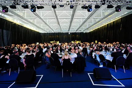 Large banquet hall with numerous guests seated at round tables, dressed formally for an event with candles and dim lighting.