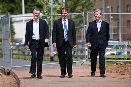 Three men in business suits walking side by side on a paved path outdoors with construction fencing in the background.