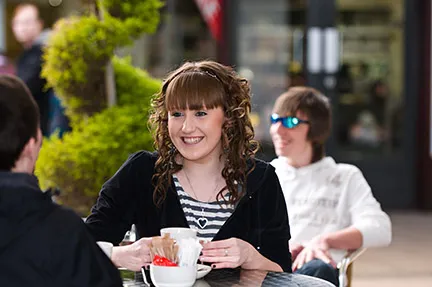Young woman with curly hair smiling while holding a coffee cup at an outdoor café table with two friends.