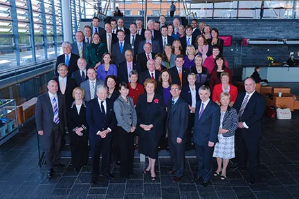 Group of professionally dressed men and women posing for a formal photo inside a modern building with large windows and stairs.