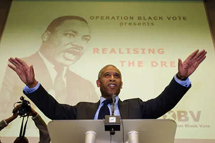 A man in a suit stands at a podium with arms outstretched, speaking in front of a projected image of Martin Luther King Jr. and text about Operation Black Vote.