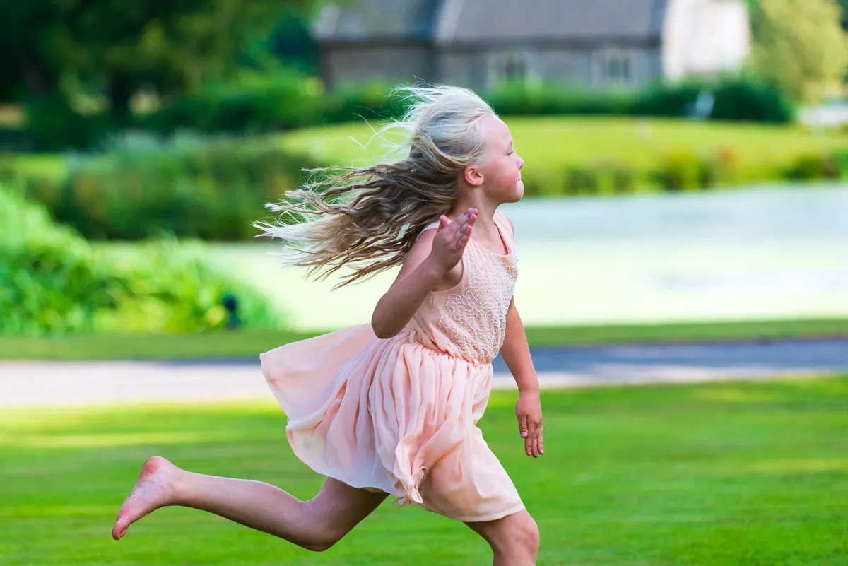 Young girl with flowing hair in a pink dress running on green grass near a pond.