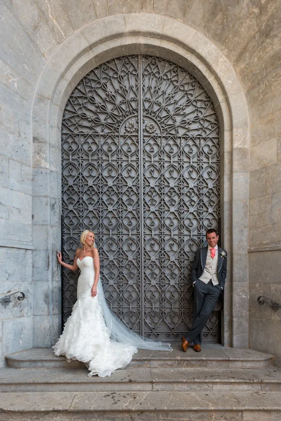 Bride in a white wedding gown and groom in a suit with a coral tie stand on stone steps in front of a large ornate wrought iron door.