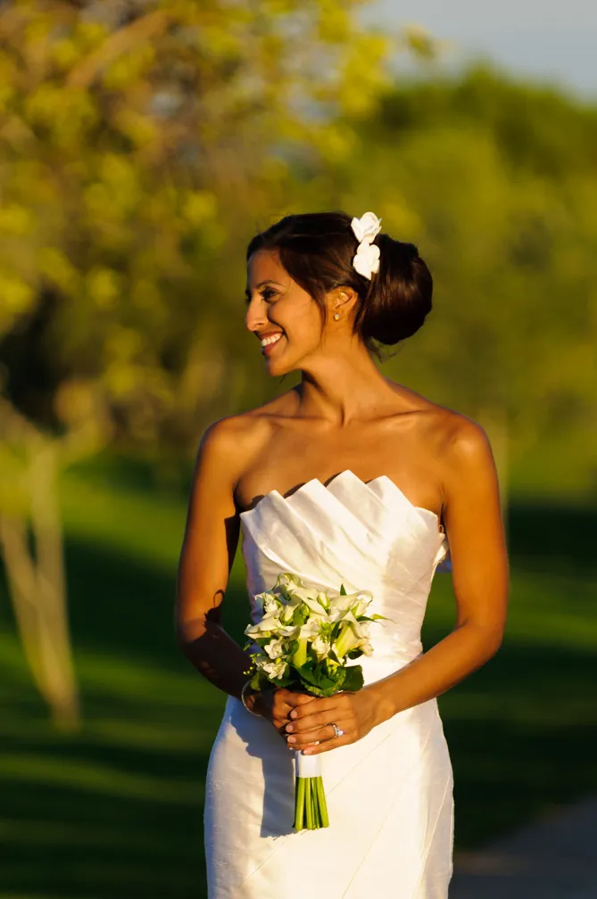 Bride in a strapless white wedding dress holding a bouquet of white flowers, smiling and looking to the side outdoors.