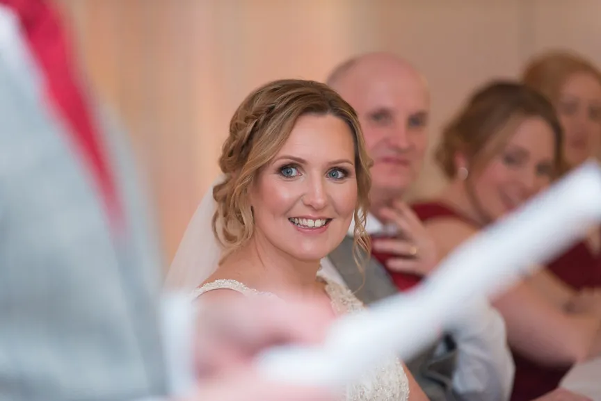 Smiling bride in a white wedding dress with braided hair looks towards a person holding a paper, with guests blurred in the background.