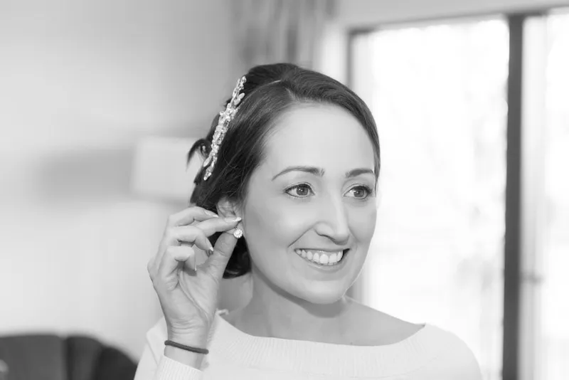 Smiling woman with hair accessory putting on an earring indoors near a window.