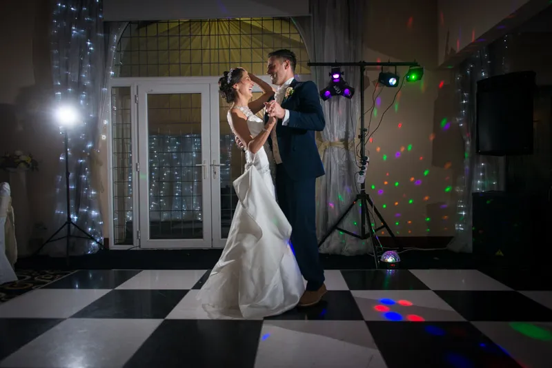 Bride and groom dancing on a black and white checkered floor with colorful party lights in the background.