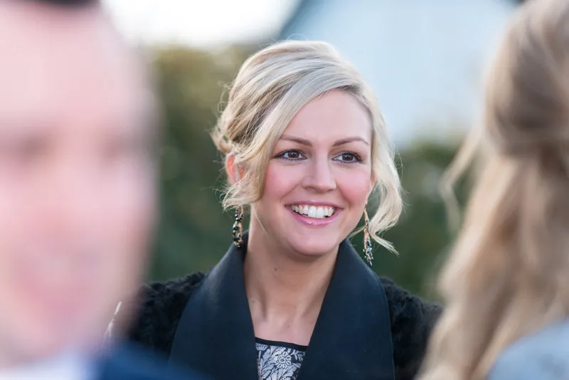 Smiling blonde woman with earrings and a black coat talking outdoors with blurred people around her.