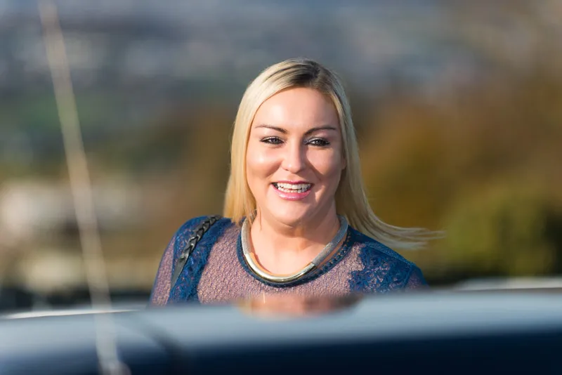 Smiling blonde woman in a blue lace top and silver necklace standing outdoors near a car.
