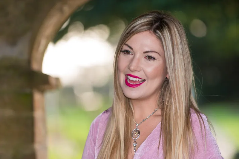Smiling woman with long blonde hair wearing a pink top and a silver pendant necklace outdoors.