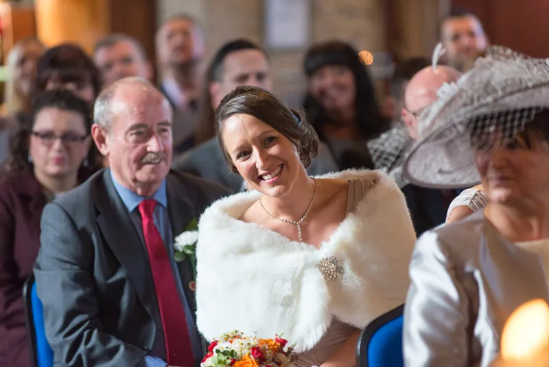 Smiling bride wearing a white fur shawl seated among guests in a wedding ceremony.