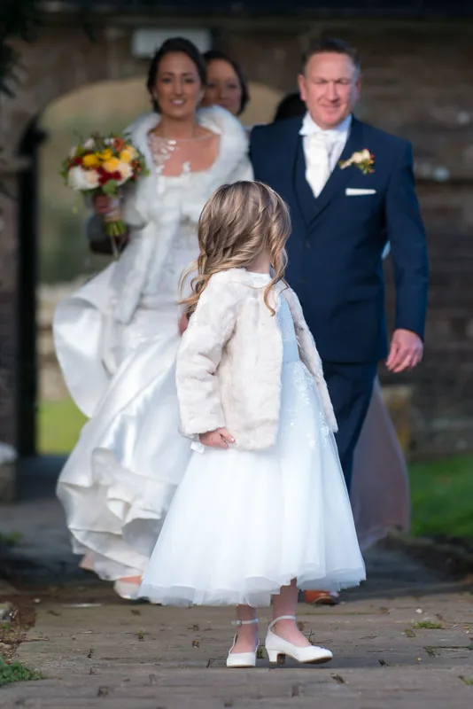 Young girl in a white dress and light jacket standing on a stone path with a bride holding a bouquet and a man in a blue suit blurred in the background.