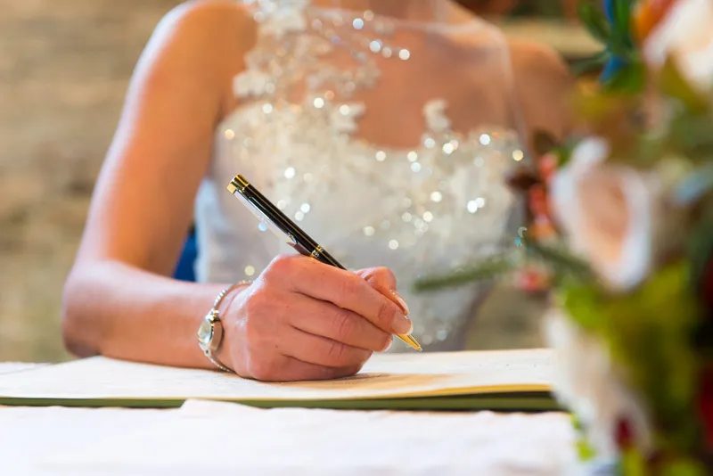 Bride in a white wedding dress signing a document with a pen, with blurred flowers in the foreground.
