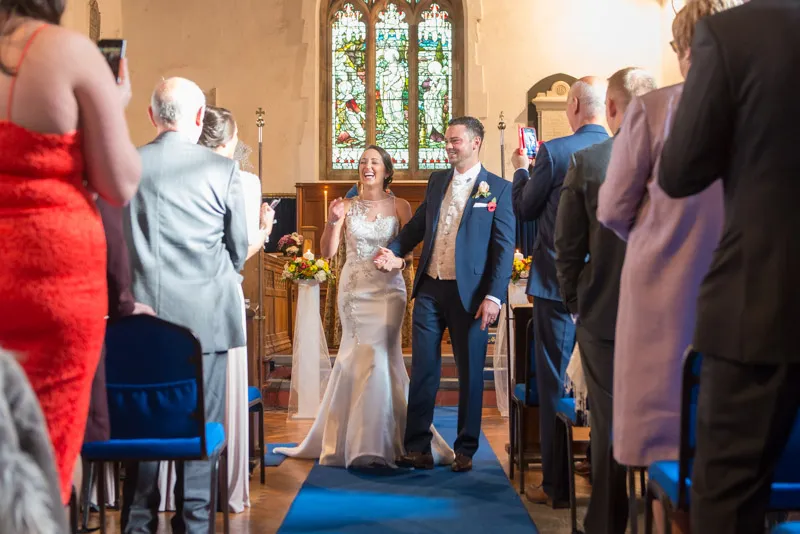 Newlywed bride and groom holding hands and smiling as they walk down the aisle of a church with guests standing on either side.
