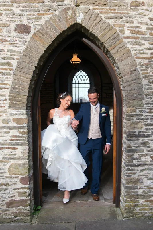 Bride and groom holding hands and smiling as they walk through a stone arched doorway.