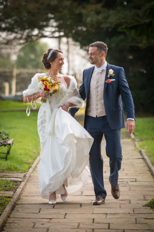 Bride in white gown and fur stole holding bouquet walks arm-in-arm with groom in navy suit on stone path outdoors.