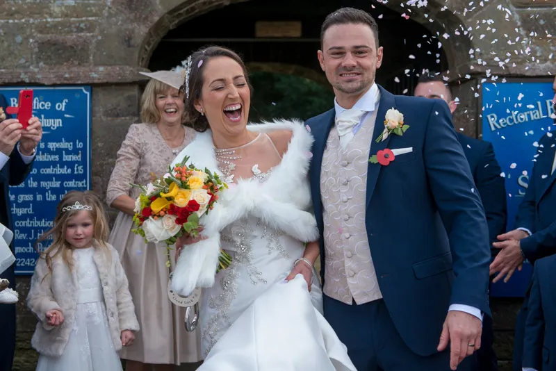 Bride in a white wedding gown and groom in a blue suit smiling and walking with guests throwing confetti.