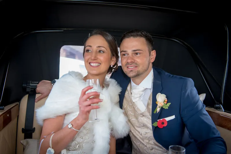 Smiling bride in white fur wrap and groom in blue suit sitting together inside a vintage car.