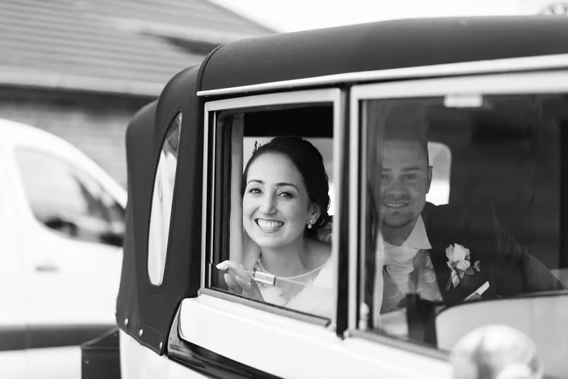 Smiling bride and groom sitting inside a vintage car, with the bride leaning out of the window.