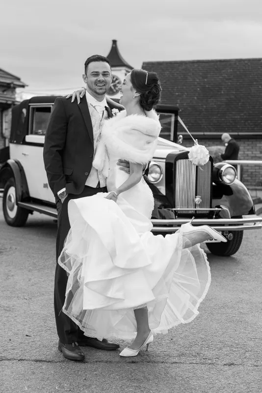 Black and white photo of a bride and groom standing in front of a vintage car, the bride lifting her dress and smiling at the groom.