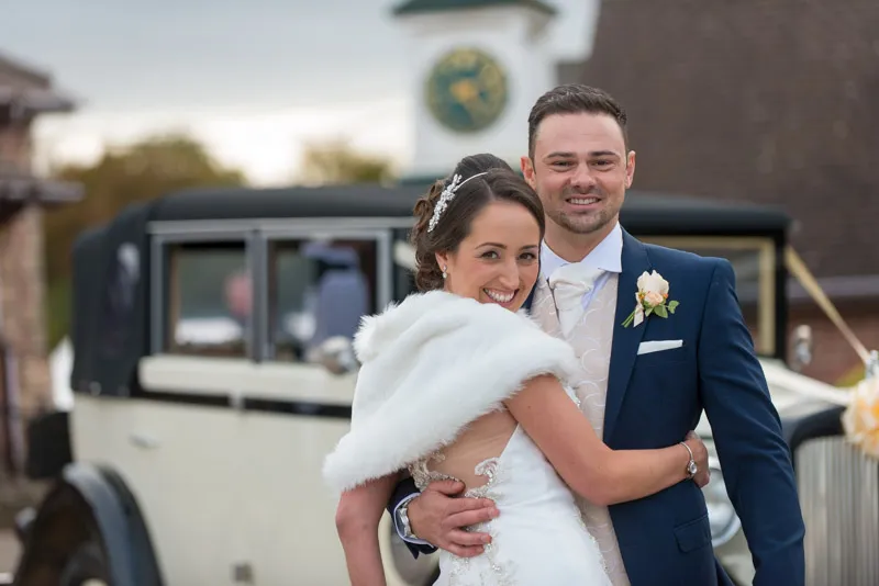 Bride in white dress and fur shawl embracing groom in navy suit with vintage car in the background.