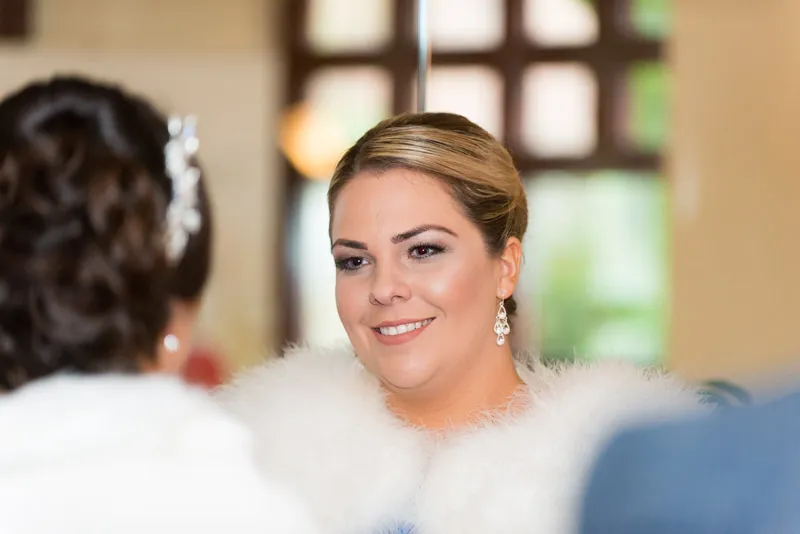 Smiling woman with styled hair and dangling earrings wearing a white fur shawl, facing a mirror indoors.