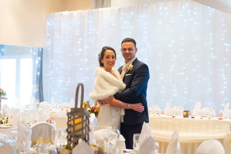 Bride wearing a white fur stole and groom in dark suit embracing in a decorated wedding reception hall with white drapes and fairy lights.