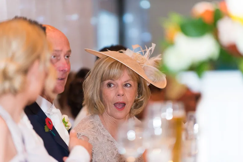 Woman in a beige hat with feathers and lace dress showing surprise, seated among guests at a formal event.