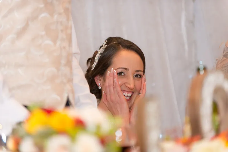 Smiling bride with hands on her cheeks, wearing a jeweled hair accessory and pearl earrings at a wedding reception.