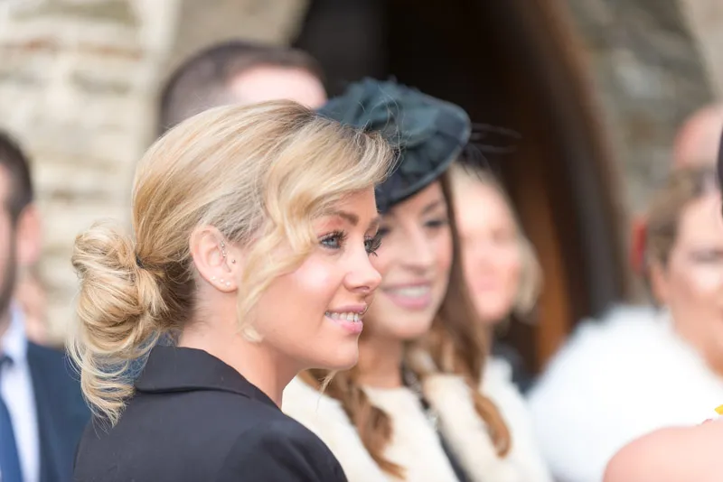 Blonde woman with a low bun hairstyle and black blazer smiling, with other people blurred in the background.