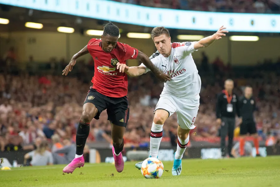 Two soccer players competing for the ball during a match, one in a red Manchester United jersey and the other in a white AC Milan jersey.