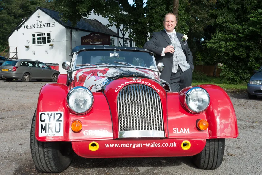 Man in a tuxedo standing beside a red vintage Morgan car with a 'Champions' design on the hood in front of The Open Hearth restaurant.