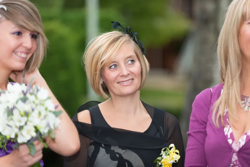 Woman with short blonde hair wearing a black dress with a floral accessory, smiling and looking sideways between two other women.