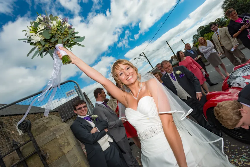 Smiling bride in a white wedding dress holding up her bouquet outdoors with guests and a vintage red car in the background under a partly cloudy sky.