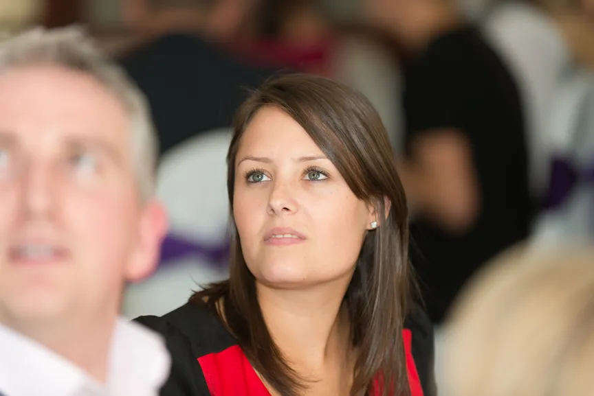 Woman with brown hair wearing a red and black top looking attentively to the side in a crowded indoor setting.