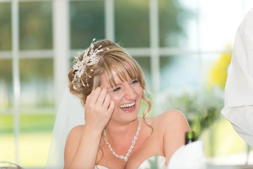 Smiling bride with decorative hairpiece and necklace sitting indoors near a window.