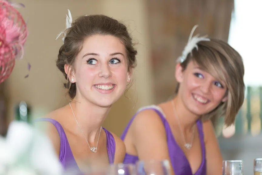 Two young women smiling and wearing purple dresses with feather hair accessories at an event.