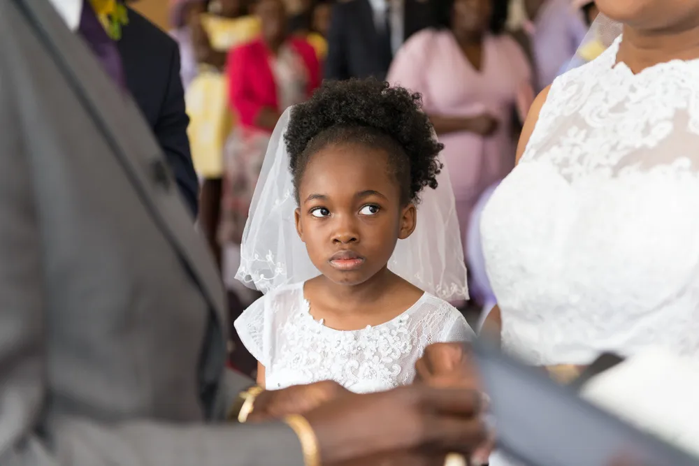 Young girl in a white lace dress and veil standing attentively between a bride and groom during a wedding ceremony.