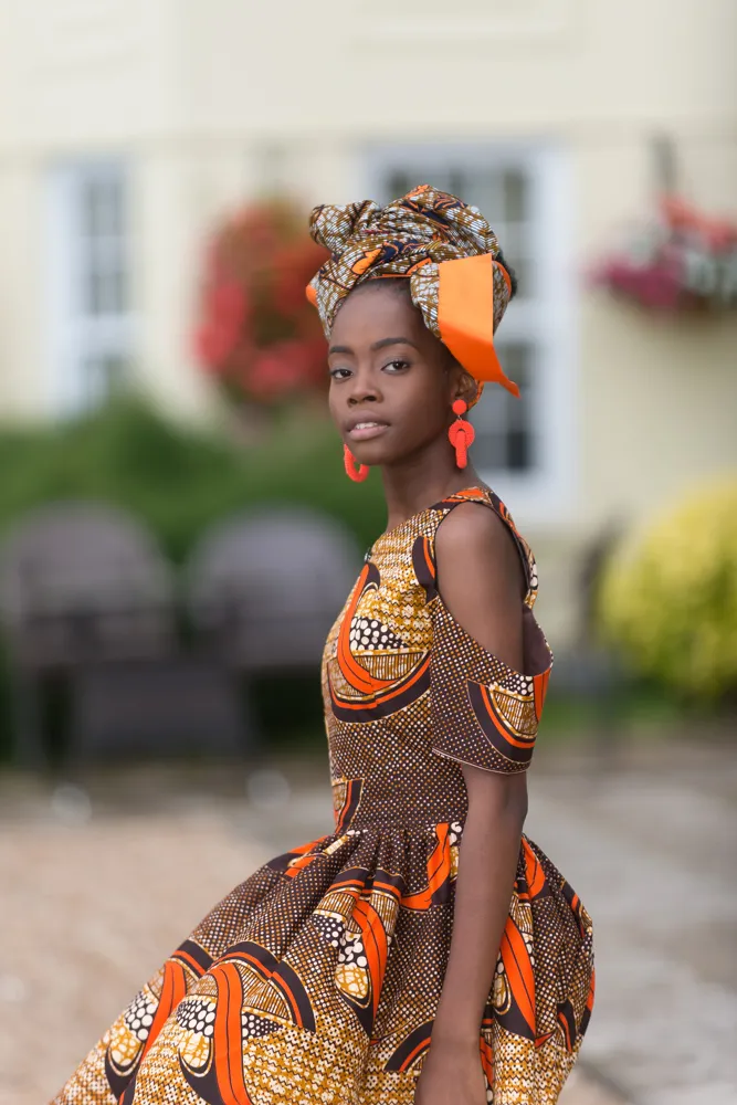 Woman wearing a colorful patterned dress and matching headwrap with orange accents, standing outdoors near blurred garden furniture and flowers.