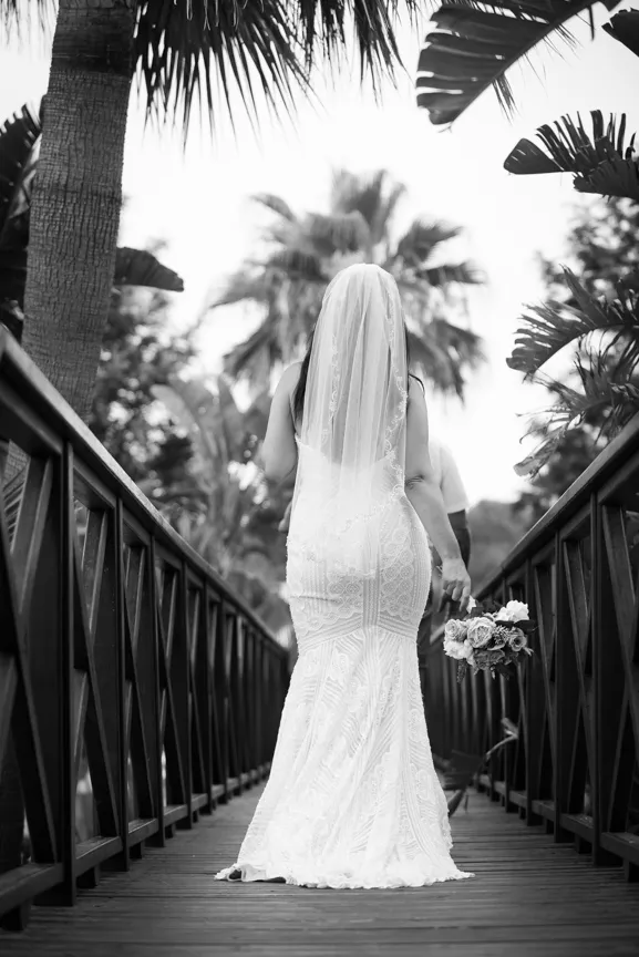 Bride in a lace wedding dress and veil holding a bouquet, walking on a wooden bridge surrounded by tropical plants.