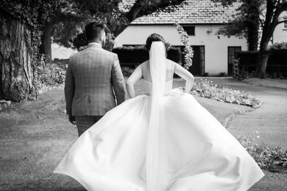 Bride in a flowing wedding dress and veil walking hand in hand with groom in a checked suit in a garden setting.