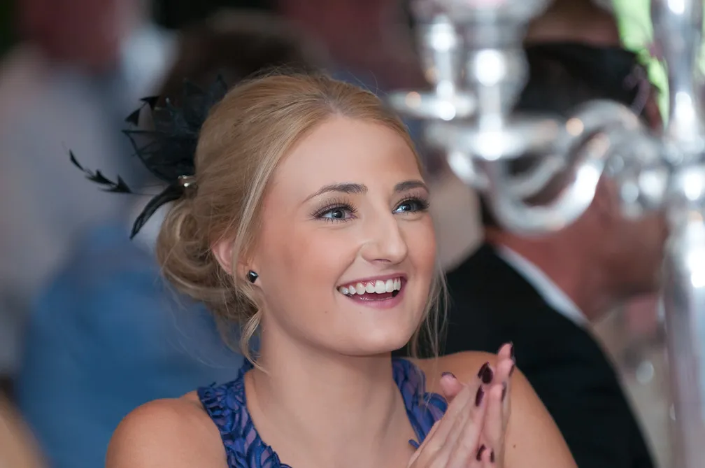 Smiling woman with blonde hair tied back adorned with a black hair accessory, wearing a blue dress, clapping hands at an indoor event.